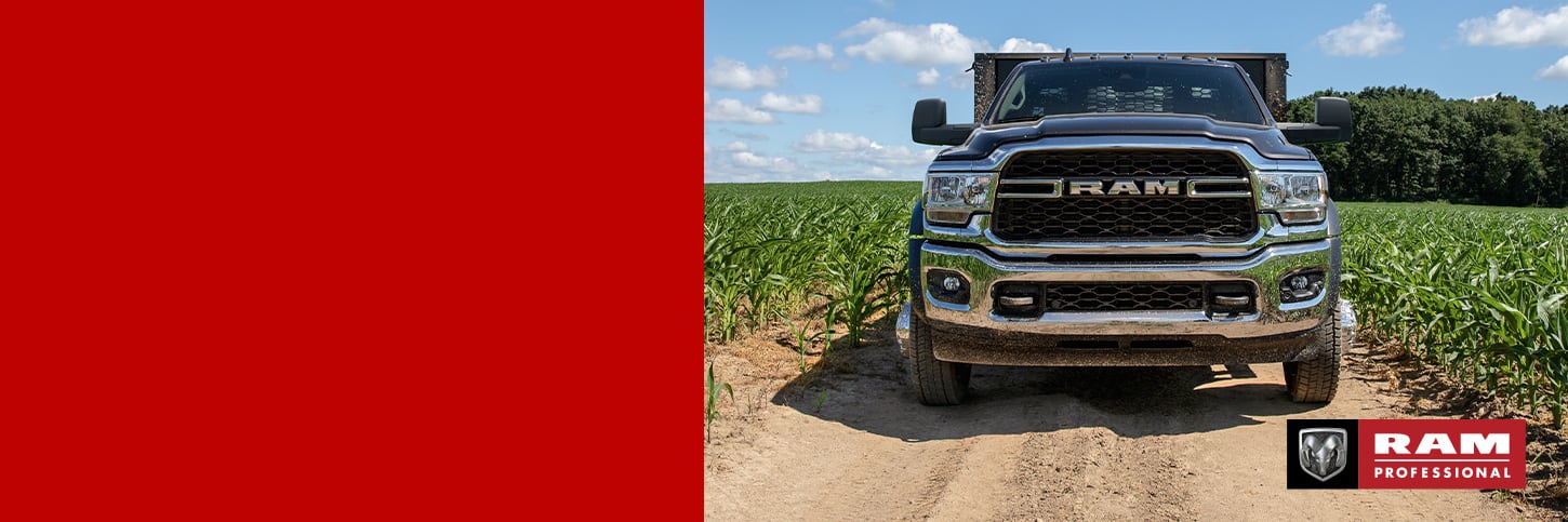 A head-on angle of a 2024 Ram 4500 Tradesman Chassis Cab with a stake bed upfit on a dirt road between fields of crops. Ram Professional.