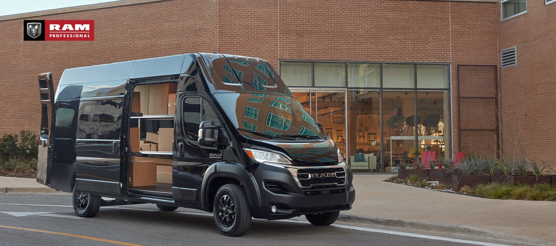 A black 2023 Ram ProMaster 3500 Super High Roof Cargo Van parked next to a furniture store, its rear doors and passenger-side sliding door open, revealing shelves full of boxes. Ram Professional.